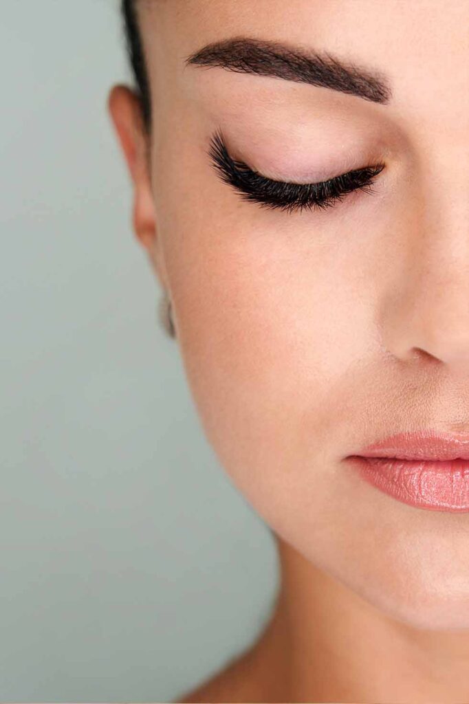 Close-up af kvindes ansigt med lukkede øjne, fremhævet af fyldige vipper og naturlige læber mod en blød grøn baggrund.