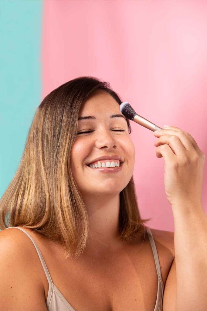 Kvinde smiler mens hun påfører makeup med en børste på kinden, foran en baggrund med lyserøde og turkise farver.